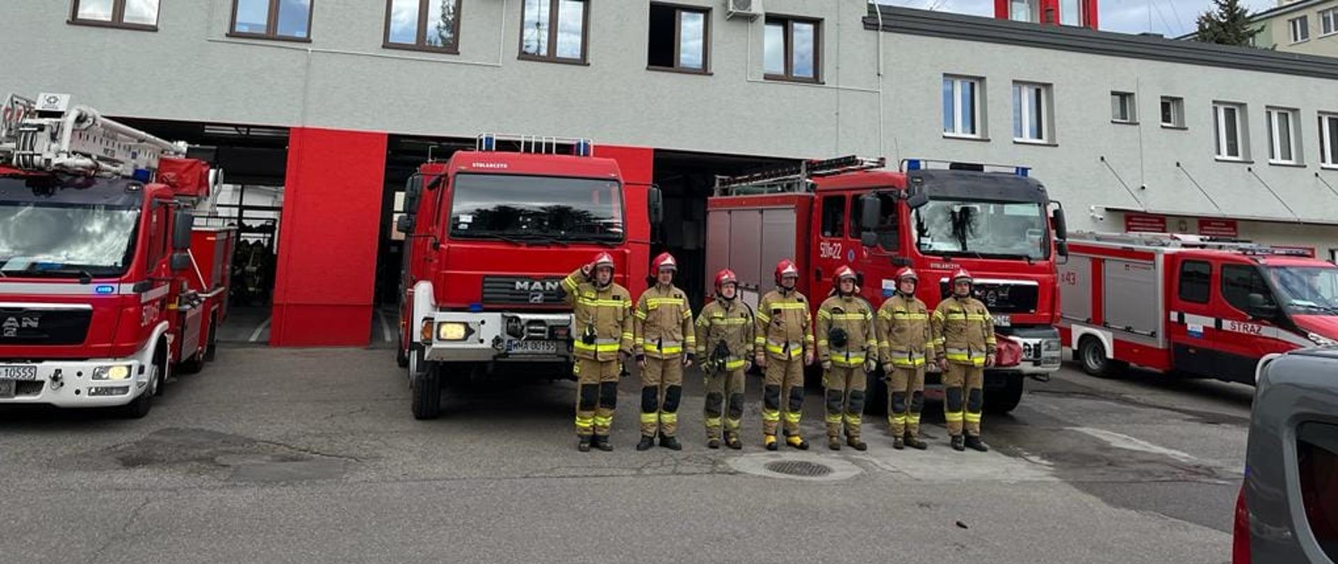 Strażacy stoją w szeregu przed samochodami strażackimi na placu Komendy. Pierwszy od lewej oddaje honor.