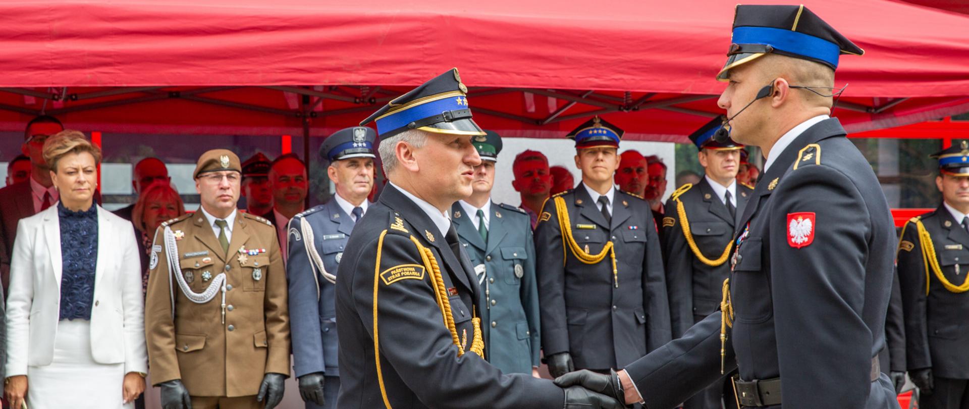 Zastępca Mazowieckiego Komendanta Wojewódzkiego PSP st. bryg. Waldemar Wysowski (w centrum, z profilu) przyjmuje meldunek o zakończeniu uroczystości z okazji Dnia Strażaka w Siedlcach. W tle, pod czerwonym namiotem ogrodowym, stoi grupa zaproszonych gości, w tym kobieta w białym żakiecie, żołnierz w oliwkowym mundurze, policjant oraz pozostali strażacy w umundurowaniu galowym.