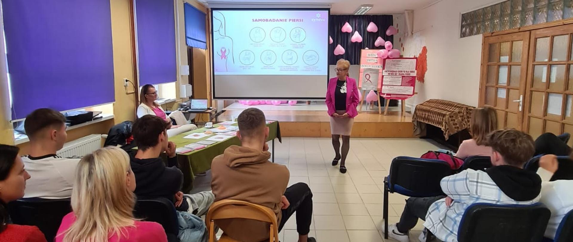 W centralnej części zdjęcia znajduje się kobieta, która przemawia do zgromadzonych gości, w tle jest przedstawiana prezentacja 