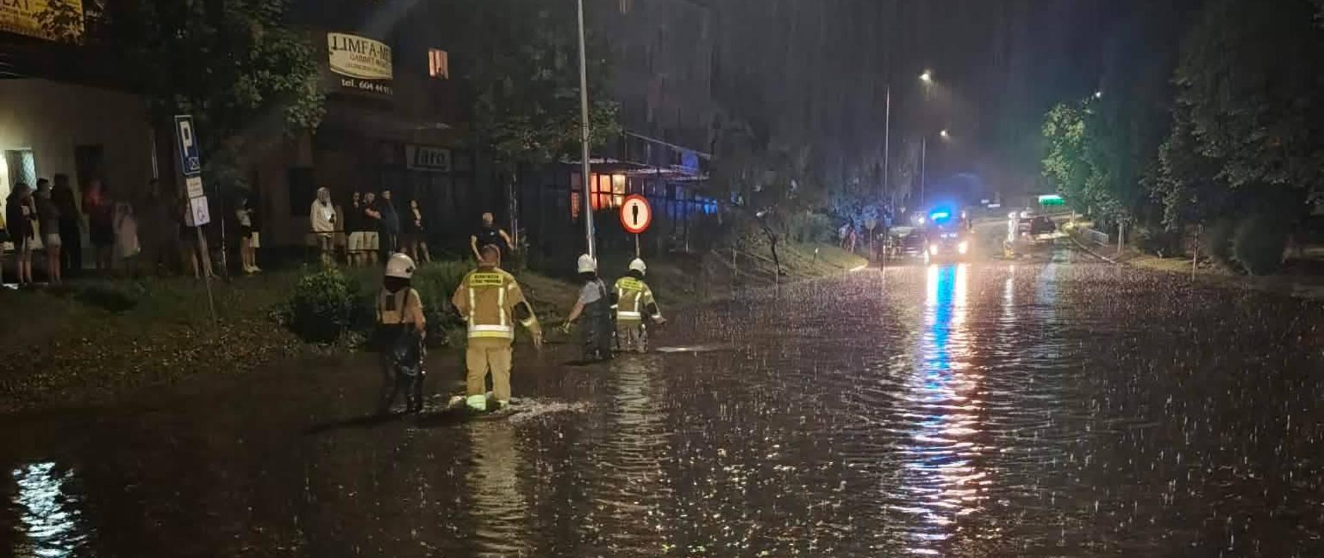 Noc, zalana droga. Strażacy w żółtych ubraniach. Z lewej strony tłum gapiów. Po drugiej stronie Samochód bojowy PSP z błyskającymi niebieskimi światłami