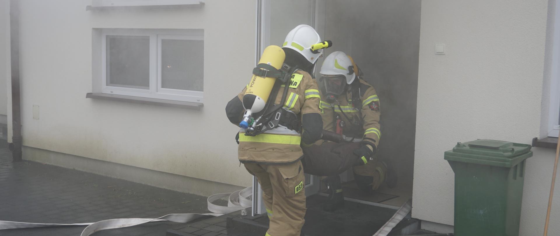 Zdjęcie przedstawia strażaków OSP wynoszących osobę poszkodowaną z zadymionego budynku. Strażacy pracują w aparatach ochrony dróg oddechowych.