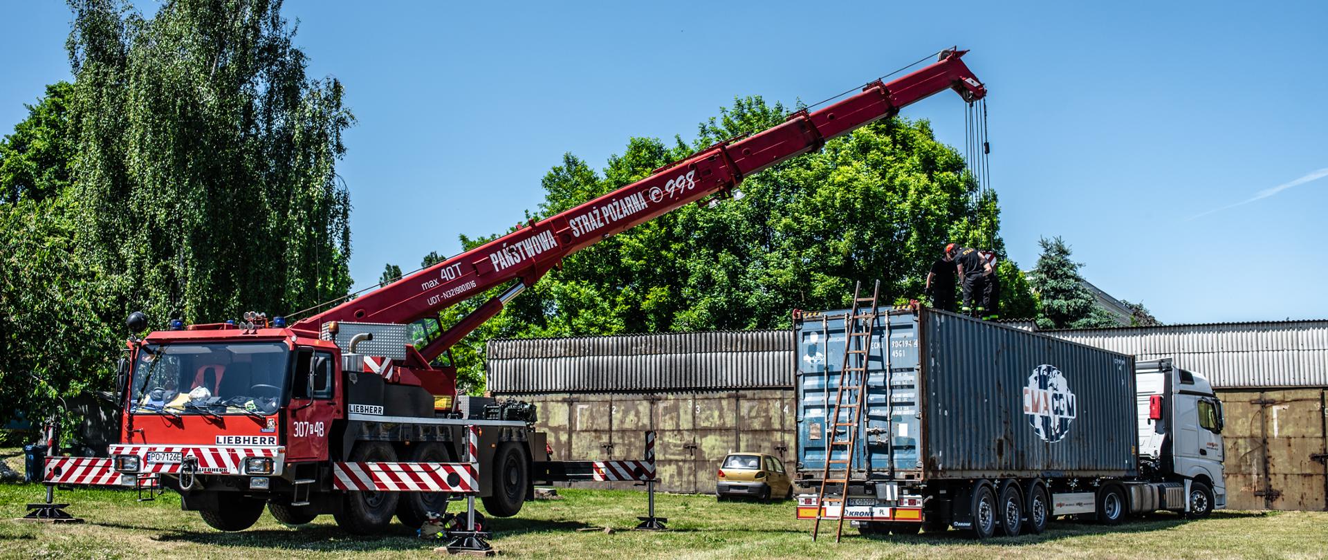 Po lewej stronie zdjęcia samochód dźwigowy w kolorze czerwonym. Pojazd jezd oznakowany numerami operacyjnymi. Posiada sygnalizację ostrzegawczą. Ramie dźwigu wysunięte nad samochód na którym znajduje się kontener morski. Na kontenerze strażacy z SGRT mocują liny do mocowań. Kontener jest w kolorze niebieskim. Ciągnik siodłowy jest w kolorze białym. Za dźwigiem drzewa oraz krzewy zielone. Za ciągnikiem siodłowym bramy garażowe w kolorze zielonym. Na pierwszym planie trawa, miejscami wypalona. Pomiędzy dźwigiem, a kontenerem samochód osobowy Matiz w kolorze złotym, który służy do ćwiczeń. Niebo bezchmurne, kolor niebieski.