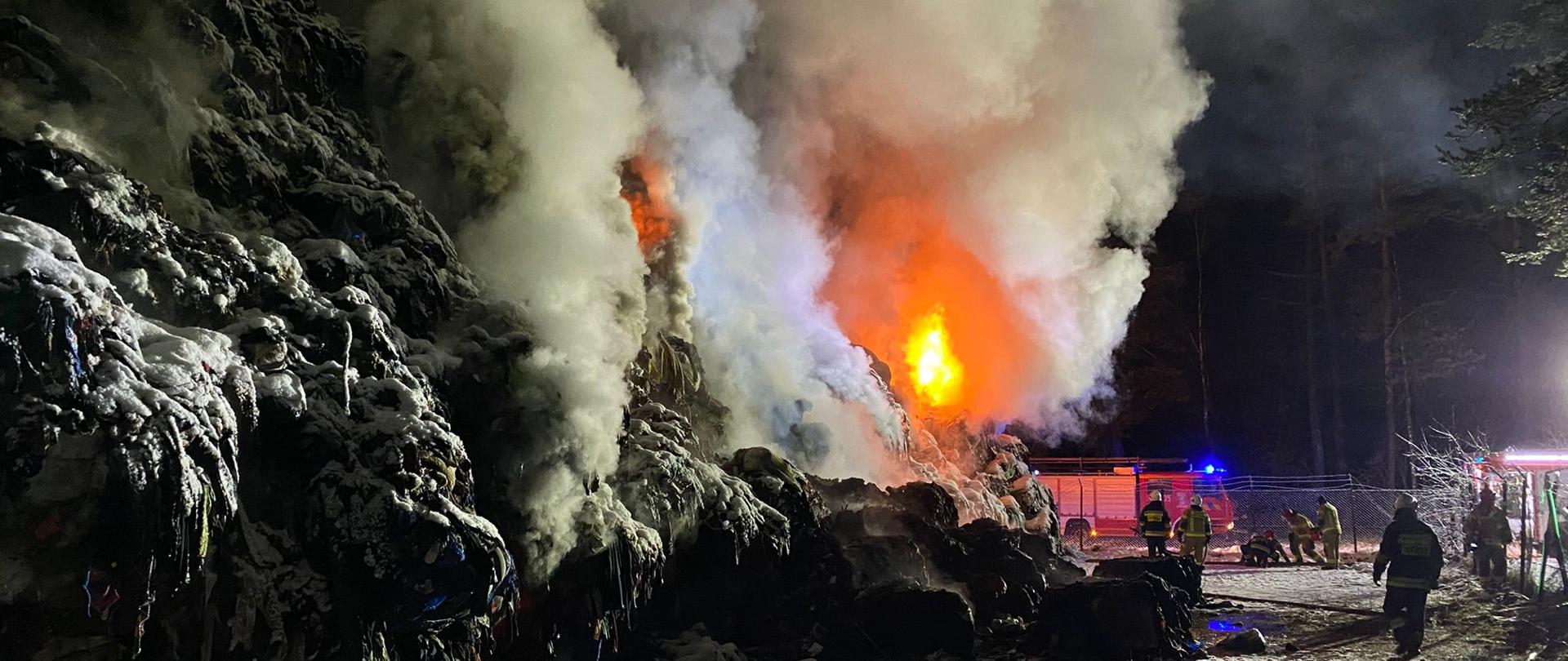 Strażacy w trakcie działań gaśniczych sprasowanych w belach materiałów tekstylnych składowanych na pryzmie. Dookoła leży śnieg. Działania prowadzone w porze nocnej. Za ogrodzeniem stoi samochód ratowniczo - gaśniczy. Po lewej stronie zdjęcia pali się wysoka pryzma materiałów tekstylnych. 