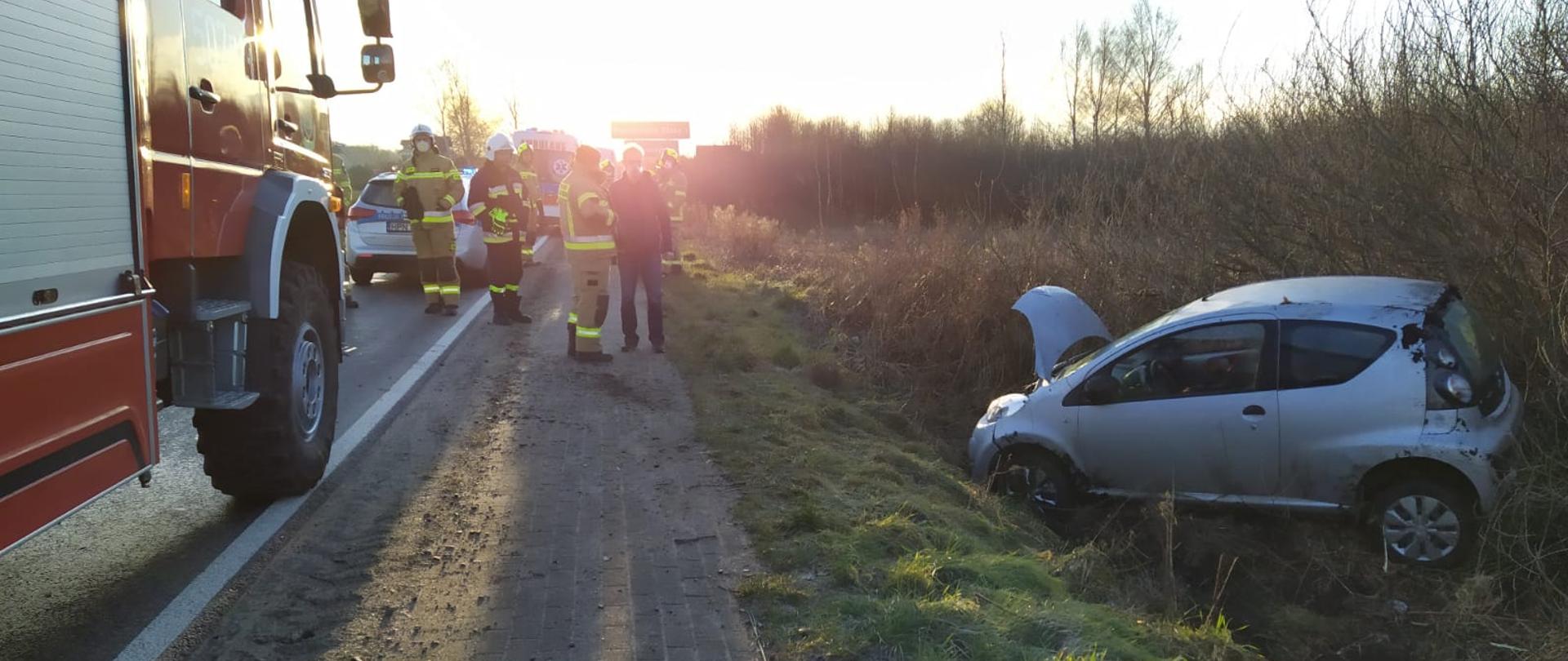 Na zdjęciu na jezdni znajdują się w tle samochody straży pożarnej oraz strażacy poza jezdnią samochód po dachowaniu