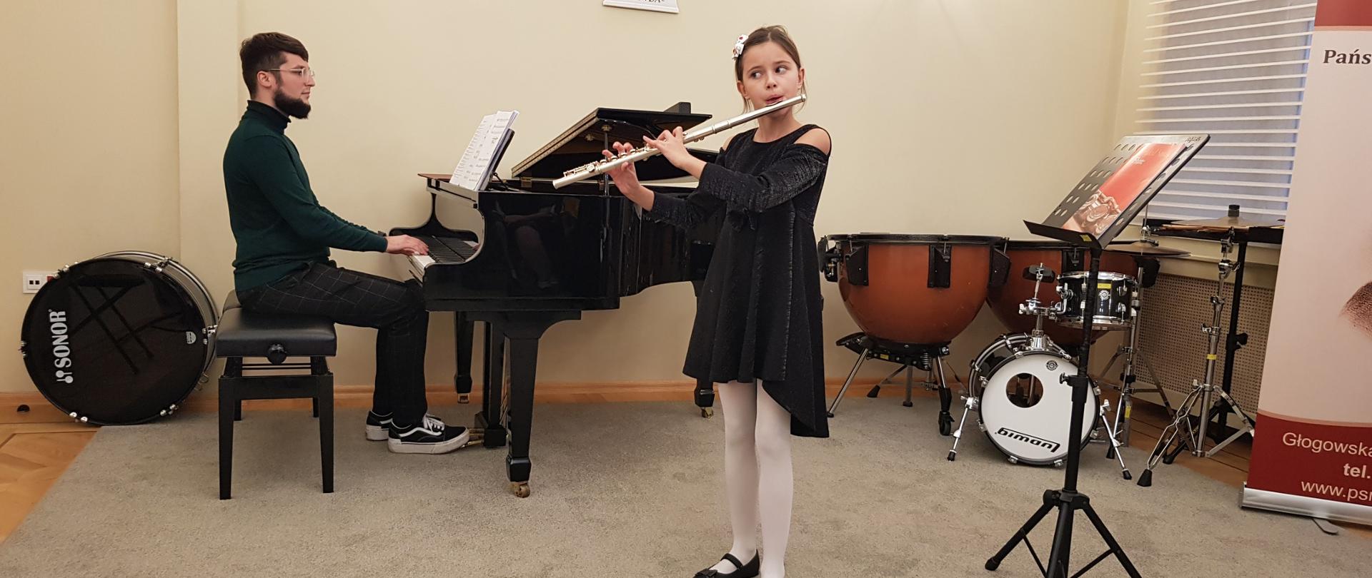 W centralnej części dziewczynka grająca na flecie. Z lewej strony akompaniator grający na fortepianie. Z prawej widoczne instrumenty perkusyjne.