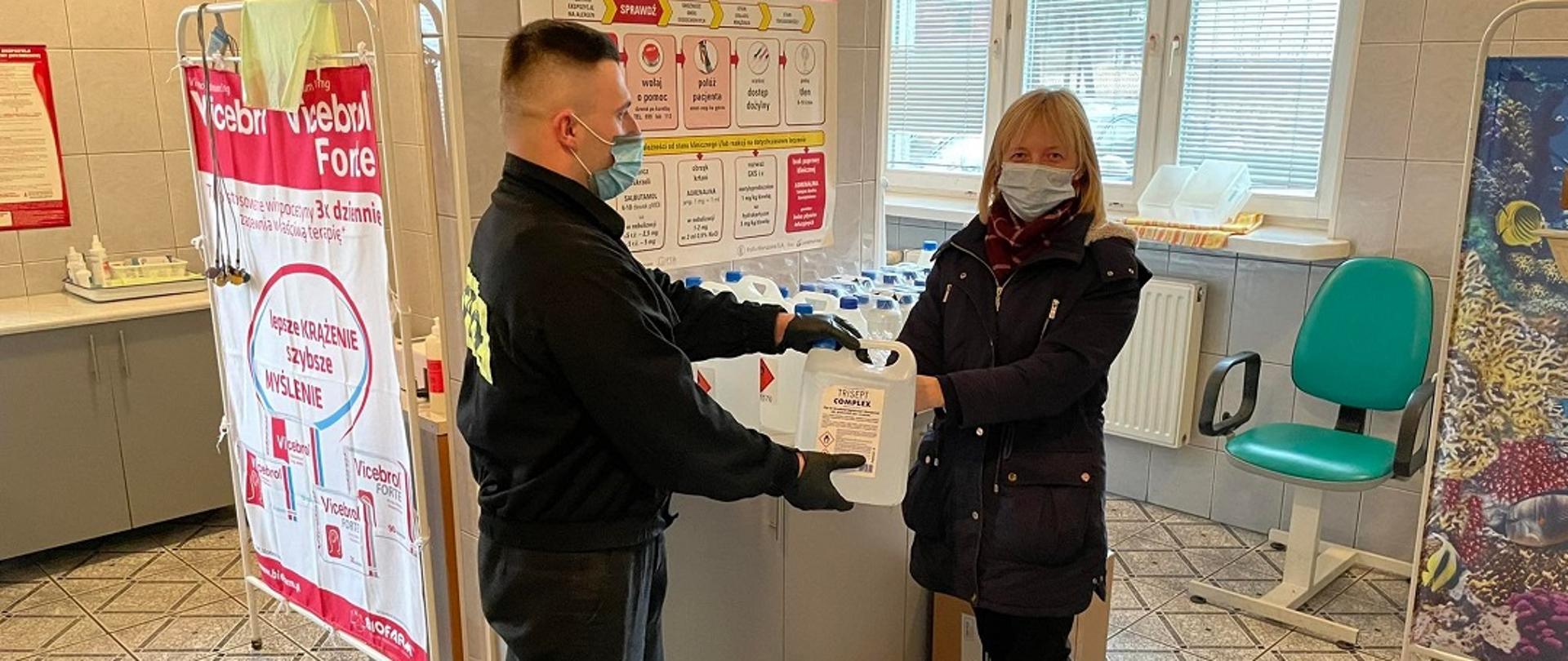 Zdjęcie obrazuje skarżyskiego strażaka podczas przekazywania środków ochrony indywidualnej oraz płynów do dezynfekcji do jednego z punktów szczepień znajdującego się na terenie powiatu skarżyskiego. Przekazane środki mają pomóc w zapewnieniu reżimu sanitarnego podczas szczepień mieszkańców powiatu skarżyskiego.