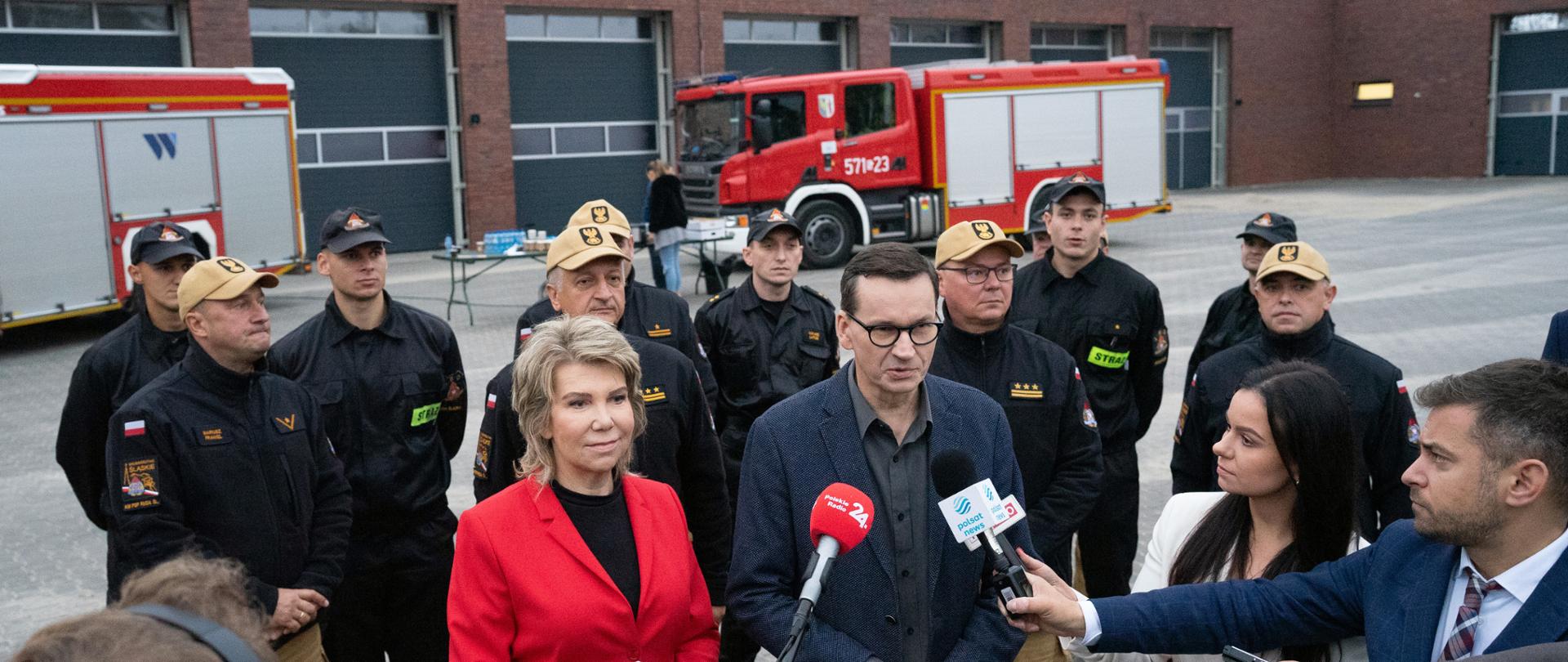 Briefing prasowy Prezesa Rady Ministrów Pana Mateusza Morawieckiego przed nową komendą Państwowej Straży Pożarnej w Rudzie Śląskiej. Premier stoi przed mikrofonami dziennikarzy , obok senator Dorota Tobiszowska. Z tyłu stoją w mundurach służbowych strażacy wśród nich zastępca śląskiego komendanta wojewódzkiego PSP st. bryg. Mirosław Synowiec i komendant miejski PSP w Rudzie Śląskiej st. bryg. Andrzej Małysiak.