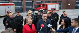 Briefing prasowy Prezesa Rady Ministrów Pana Mateusza Morawieckiego przed nową komendą Państwowej Straży Pożarnej w Rudzie Śląskiej. Premier stoi przed mikrofonami dziennikarzy , obok senator Dorota Tobiszowska. Z tyłu stoją w mundurach służbowych strażacy wśród nich zastępca śląskiego komendanta wojewódzkiego PSP st. bryg. Mirosław Synowiec i komendant miejski PSP w Rudzie Śląskiej st. bryg. Andrzej Małysiak.