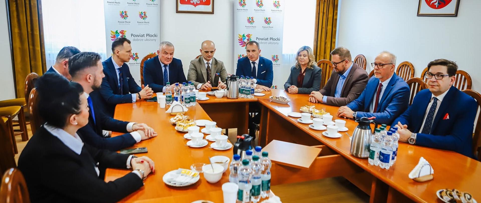 W jasnym pomieszczeniu o charakterze biurowym lub urzędowym odbywa się spotkanie oficjalne grupy jedenastu osób, siedzących przy dużym stole konferencyjnym w kształcie litery „U”. Wszyscy są ubrani w stroje formalne. Uczestnicy są zwróceni ku sobie, co sugeruje trwającą dyskusję lub naradę.