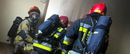 czterech strażaków ubranych w ubrania specjalne przygotowuje się do wejścia do pomieszczenia objętego pożarem, z drzwi wydobywa się dym