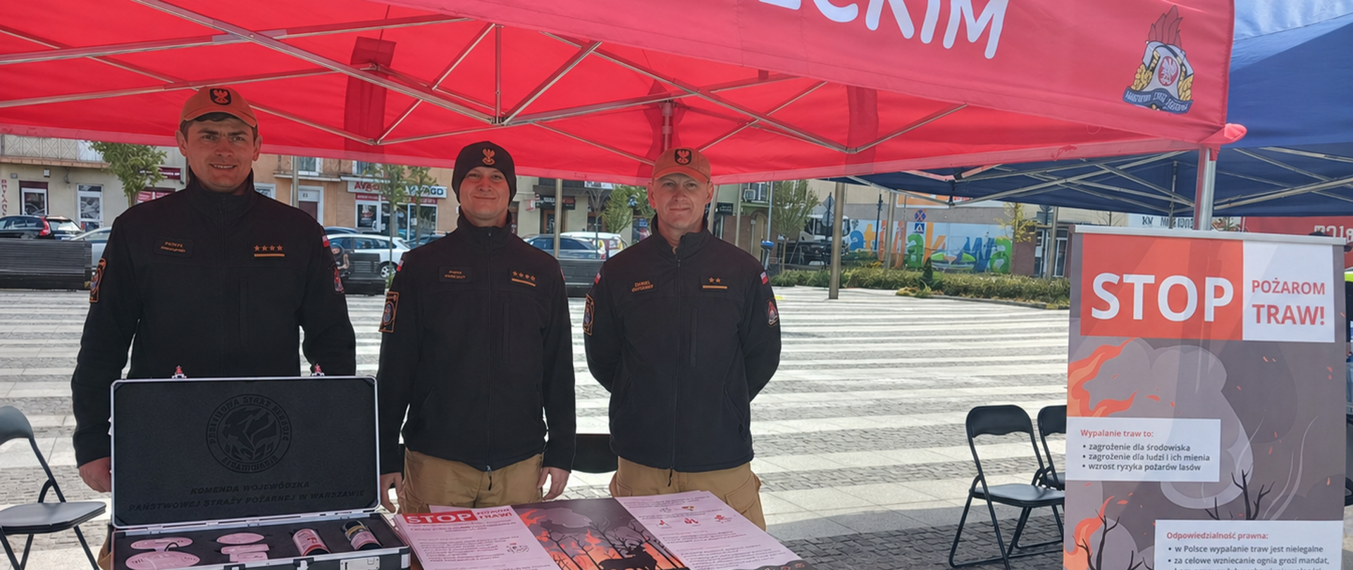 Trzech funkcjonariuszy PSP stoi przy stoisku straży pożarnej, na stoliku znajduje się walizka z czujkami oraz plakaty. Z prawej strony stoi plakat akcji Stop Pożarom traw