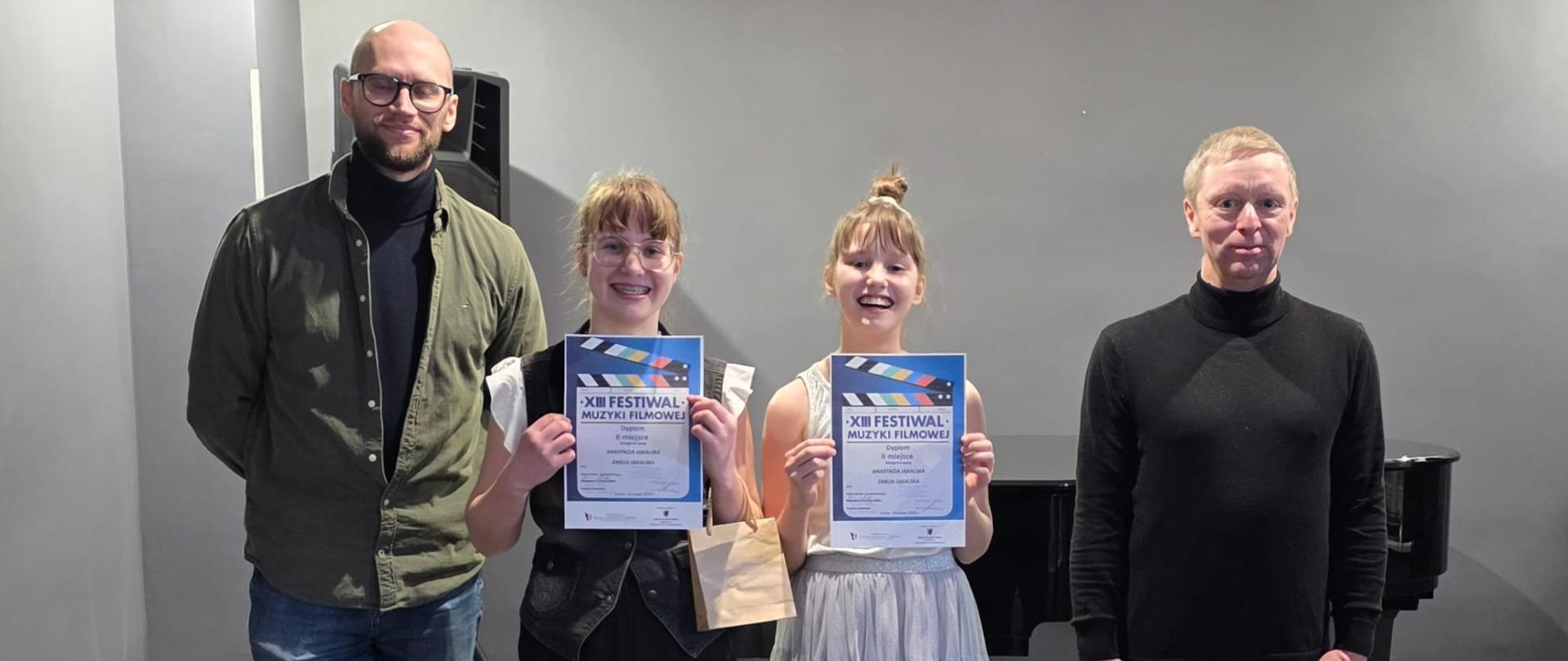 Na scenie stoją cztery osoby pozujące do zdjęcia podczas wydarzenia muzycznego.
Po lewej stronie stoi wysoki mężczyzna w okularach, zielonej koszuli i brązowych butach, obok niego stoi kobieta ubrana na czarno, trzyma dyplom i uśmiecha się do aparatu.
Następnie stoi kobieta w jasnej, eleganckiej sukni z ozdobą we włosach, trzymająca dyplom, dalej widać mężczyznę ubranego na czarno. W tle czarny fortepian.