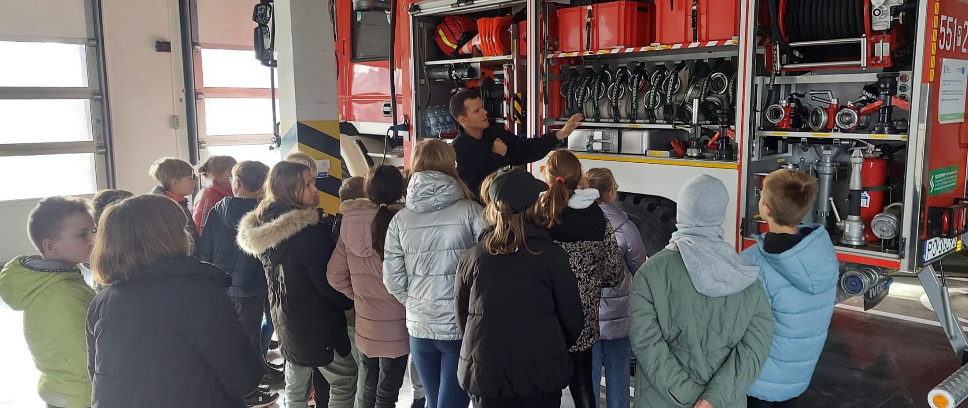 Na zdjęciu widać dzieci z Szkoły Podstawowej nr 5 w Pile oraz strażaka przedstawiającego sprzęt na samochodzie pożarniczym na garażu JRG 1.