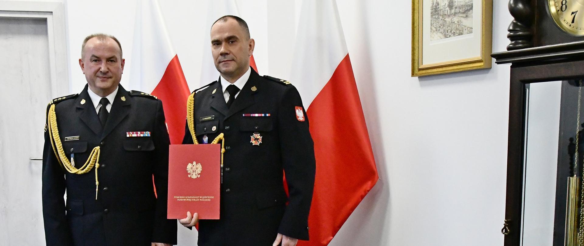 Dwóch strażaków w mundurach wyjściowych ze sznurem stoi obok siebie jeden z nich trzyma czerwoną teczkę za nimi ustawione są trzy flagi Polski są drzwi na ścianie wisi obraz a obok ustawiony jest zegar. 