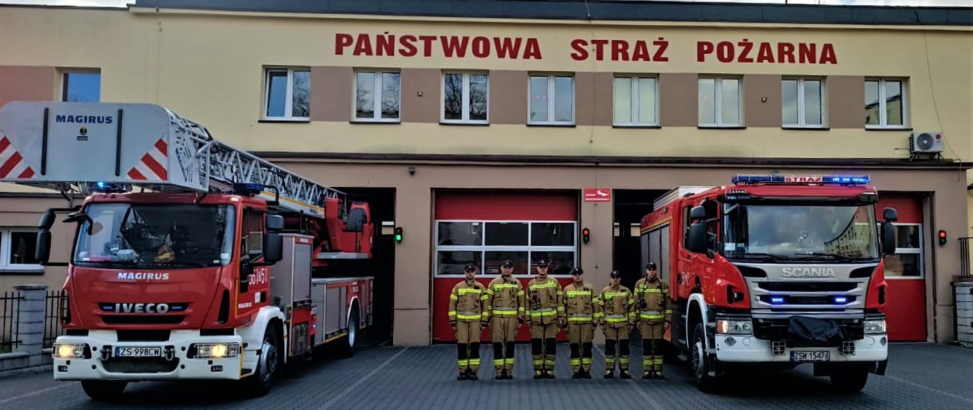 2 samochody pożarnicze - drabina mechaniczna i średni samochód ratowniczo-gaśniczy z włączonymi sygnałami świetlnymi i dźwiękowymi stoją na placu przed strażnicą. W tle strażnica z napisem na elewacji „Państwowa Straż Pożarna”. Na pierwszym planie w szeregu stoi zmiana służbowa strażaków w mundurach bojowych.