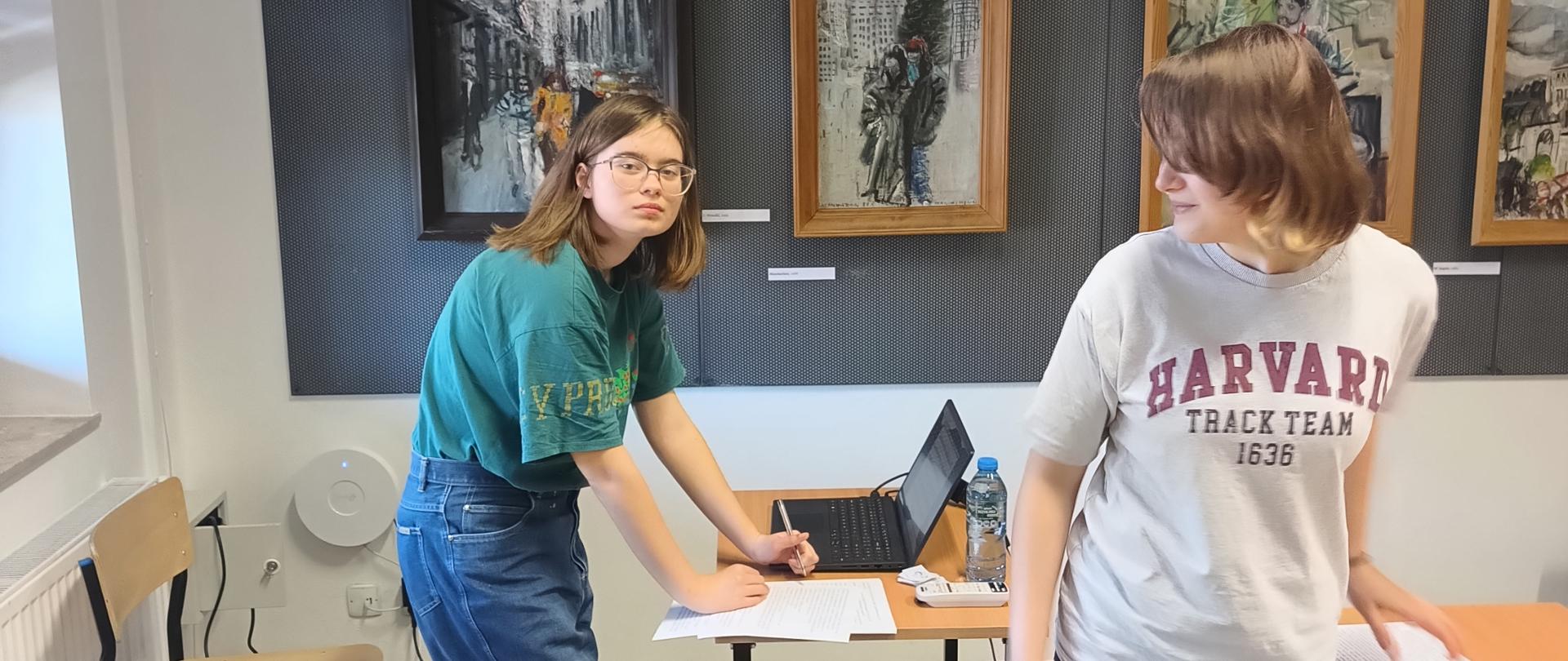 Na zdjęciu widzimy dwie uczennice: Wiktorię Białek (LO nr 3 Rzeszów) oraz Marię Mozgała (PLSP Zakopane), które w dniu 5 marca 2025 r. przeprowadziły w PLSP im. A. Kenara wykład w ramach projektu "Neuroinkluzja". Fotografia została wykonana w szkolnej galerii 'Strug". Jedna z uczennic stoi przed biurkiem na którym położony jest laptop. Uczennica w jednej ręce trzyma długopis a drugą przytrzymuje kartki papierowe położone na blacie biurka. Druga uczennica również stoi przy drugim stoliku na którym są położone materiały materiały prezentowane na wykładzie.