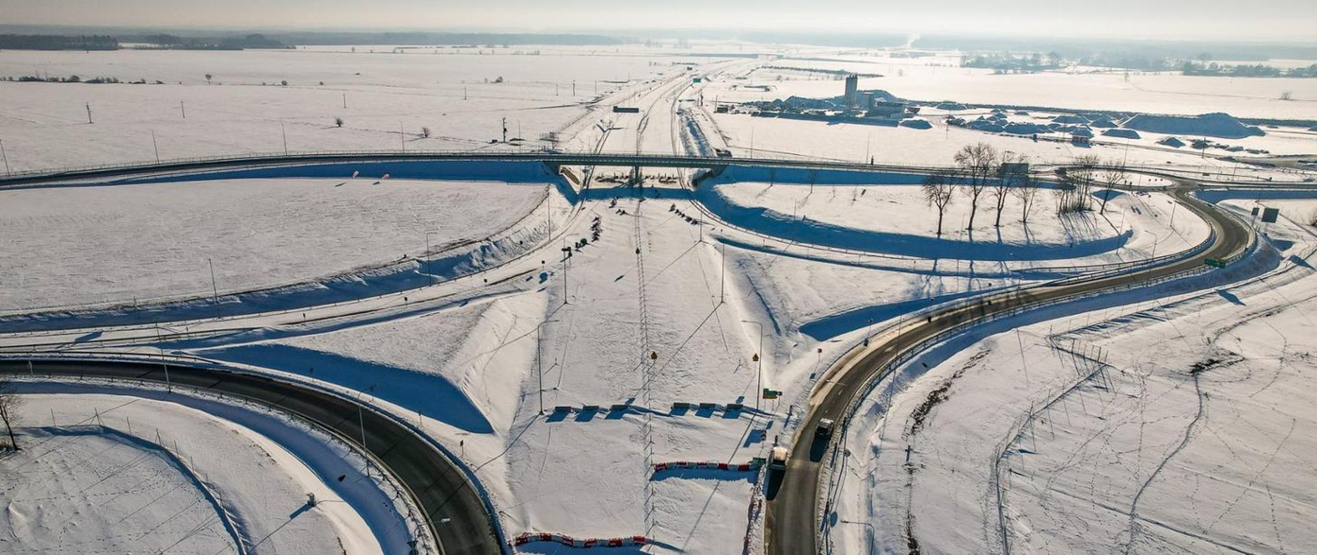 Zimowy, lotniczy widok na węzeł drogowy otoczony rozległymi, ośnieżonymi polami. Drogi łagodnie zakręcają i łączą się przy niewielkim wiadukcie, a w tle widać rozproszone zabudowania oraz szeroką perspektywę zimowego krajobrazu.