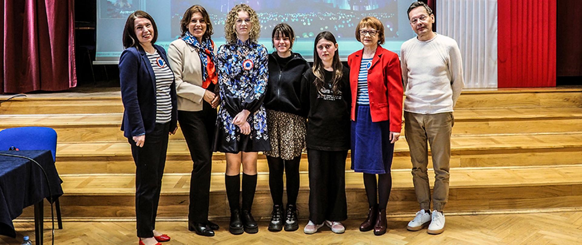Na zdjęciu znajduje się grupa siedmiu osób stojących na drewnianej podłodze w auli lub sali konferencyjnej. W tle widać duży ekran z wyświetlonym obrazem zamku nocą i fajerwerkami oraz elementy dekoracyjne, w tym biało-czerwone zasłony.
W grupie jest siedem osób, w tym czterech nauczycieli: Agnieszka Drzewiecka, Dorota Nowiszewska-Machoń, Alina Tkaczyk, Andrzej Mazuś oraz trzy uczennice.
Osoby na zdjęciu mają różnorodne stroje – od eleganckich marynarek i sukienek po bardziej casualowe ubrania. Dwie kobiety mają przypięte kokardy w barwach Francji, co może sugerować wydarzenie związane z kulturą frankofońską. Po prawej stronie grupy stoi mężczyzna ubrany w jasny sweter i spodnie, a kilka osób nosi ciemniejsze ubrania.
Niektóre osoby mają uśmiechnięte twarze, a inne bardziej poważne wyrazy twarzy. Wszystkie patrzą w stronę aparatu. Wystrój sali oraz obecność ekranu sugerują, że zdjęcie mogło zostać wykonane podczas oficjalnego wydarzenia lub prezentacji.