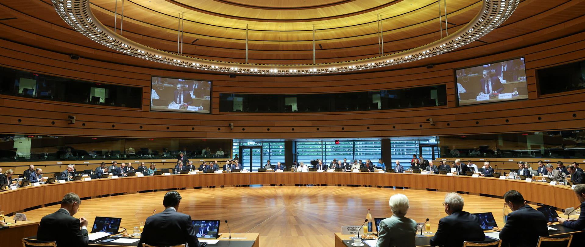 Posiedzenie Rady ECOFIN w dużej sali konferencyjnej. Uczestnicy obrad siedzą przy okrągłym układzie stołów, a przebieg spotkania jest wyświetlany na ekranach umieszczonych na ścianach sali.