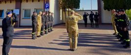 Strażacy stoją w dwóch rzędach na placu manewrowym. Na środku stoi dowództwo straży pożarnej. Na dalszym planie strażnica, maszt i poczet flagowy. Obok krzewy.