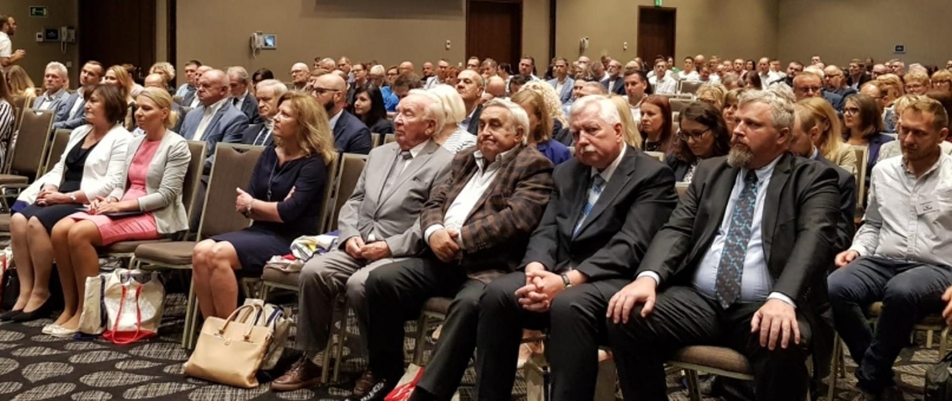 Grupa osób w strojach formalnych siedząca w sali konferencyjnej podczas wydarzenia zawodowego.