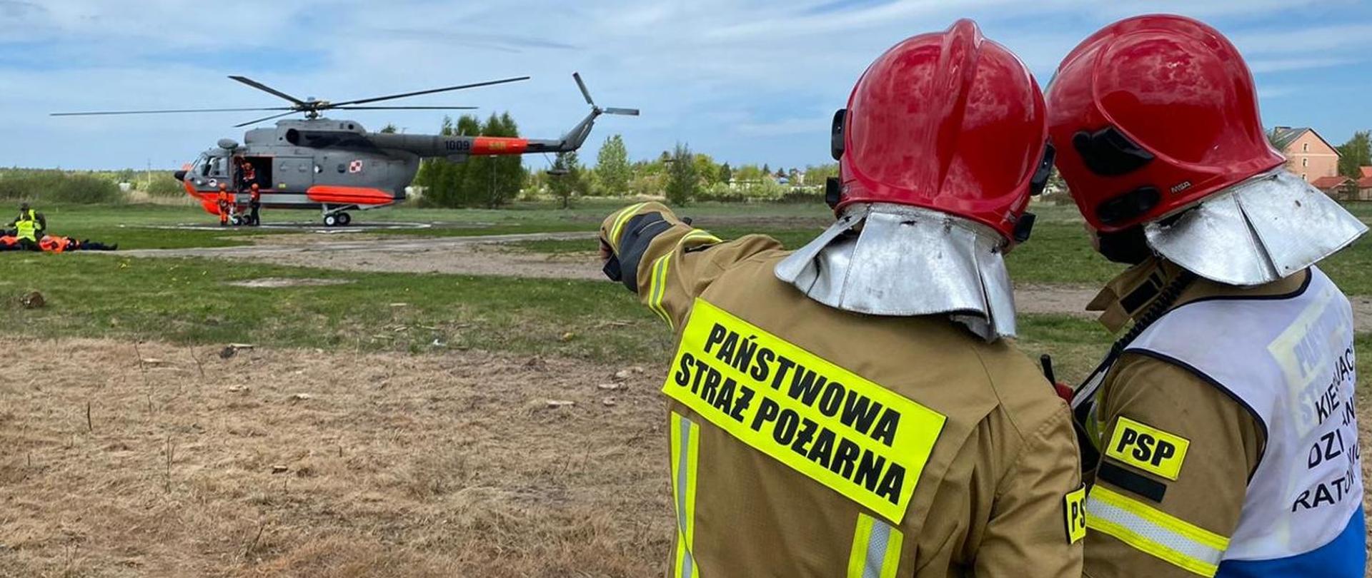 Dwóch strażaków stoi tyłem, jeden z nich wskazuje na stojący dalej helikopter
