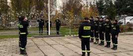 Na zdjęciu funkcjonariusze PSP w umundurowaniu koszarowym. W centralnej części maszt z flagą państwową. Zachmurzenie całkowite. Osoby stoją na betonowym, szarym podłożu. 