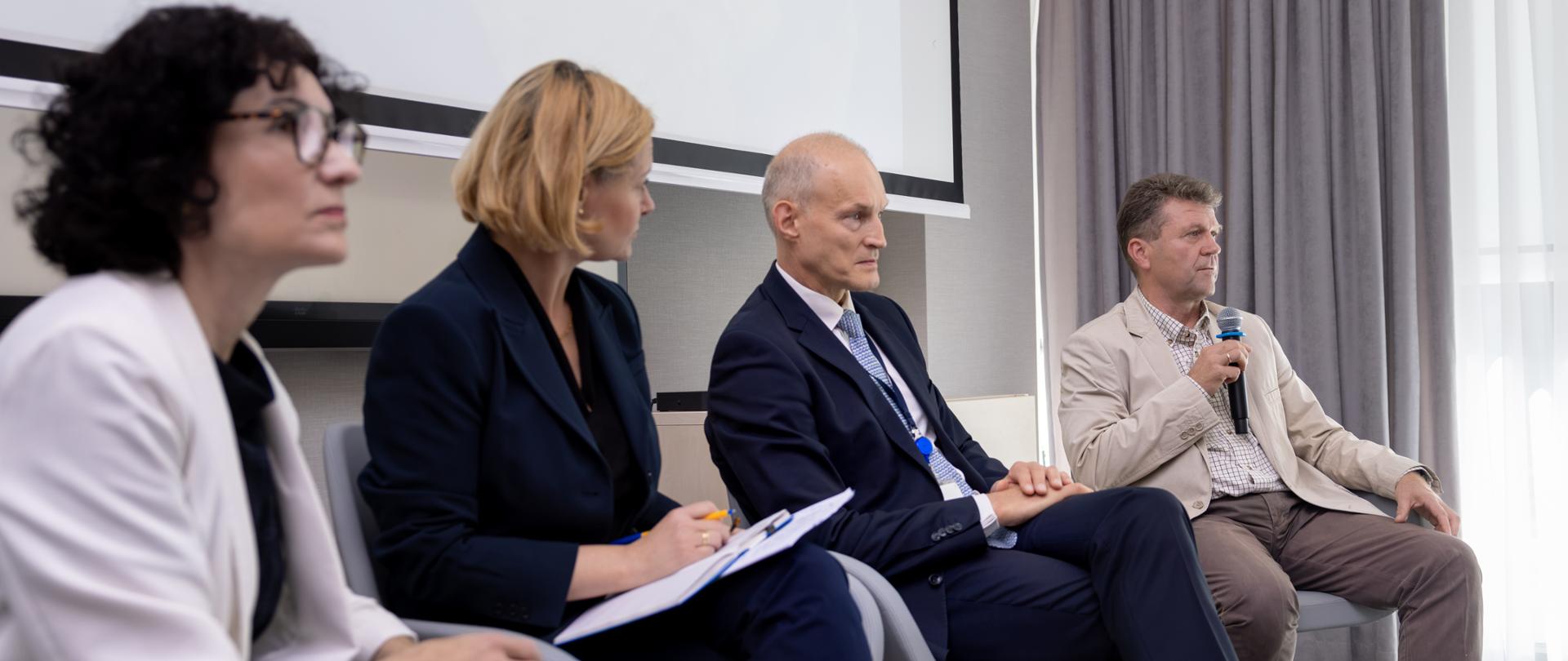 Cztery osoby uczestniczą w panelu dyskusyjnym, siedząc na fotelach w sali konferencyjnej. Po środku siedzi wiceminister Zajączkowski.