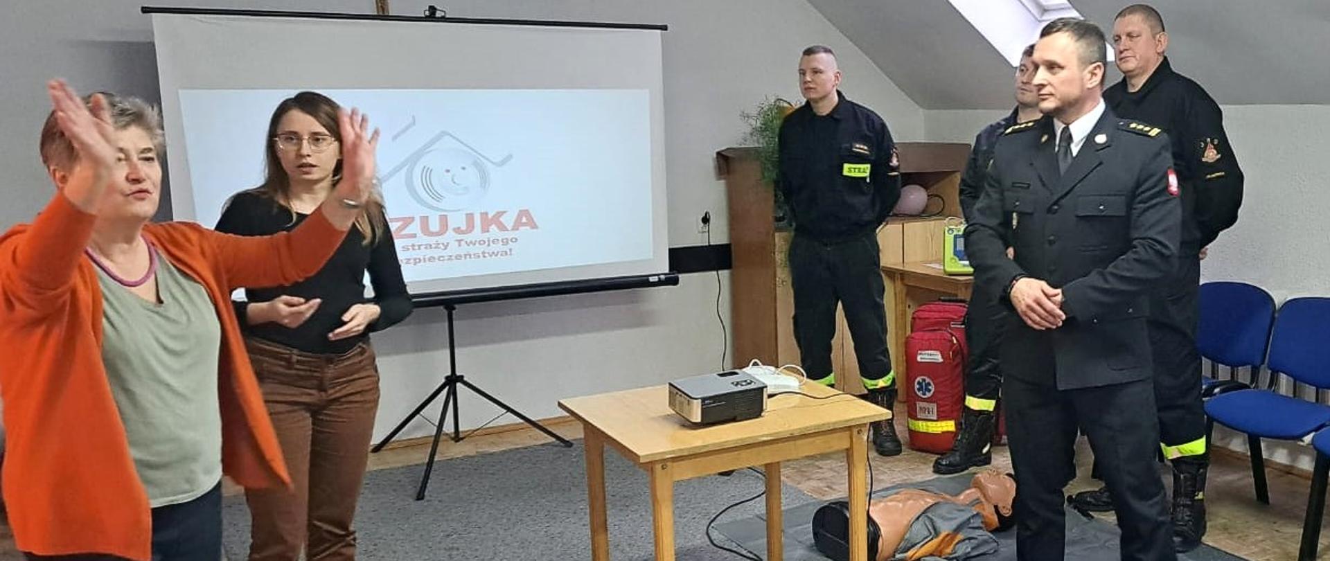 W pomieszczeniu na szarym dywanie stoi stolik. W około stoją strażacy oraz dwie kobiety.