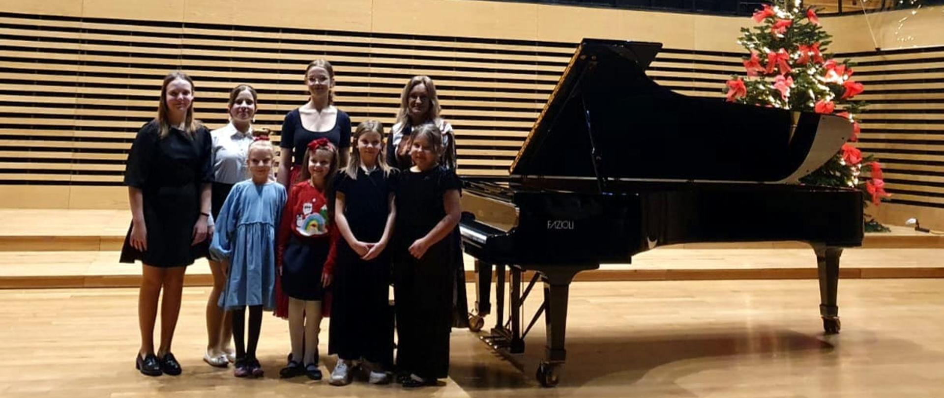 Cztery dziewczynki stoją na estradzie sali koncertowej w rzędzie, za nimi w drugim rzędzie stoją trzy nastolatki oraz kobieta. Obok po prawej stronie stoi fortepian, za nim z tyłu udekorowana choinka.