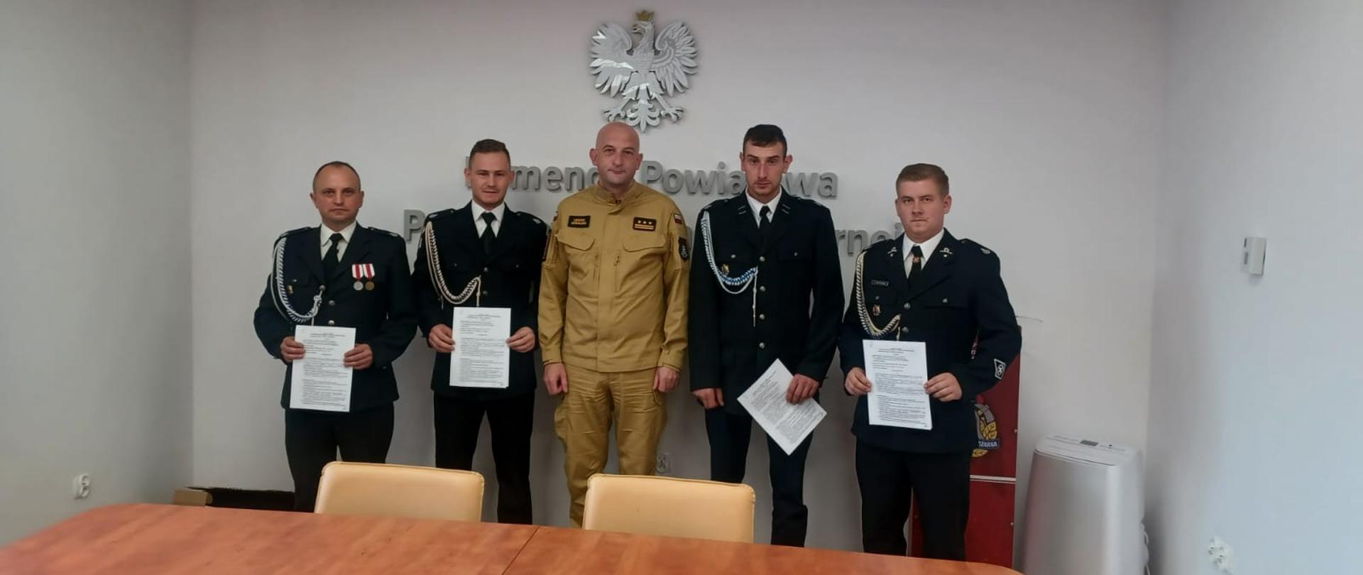 Komendant powiatowy Państwowej Straży Pożarnej w Działdowie oraz czterech strażaków-ratowników OSP pozują do zdjęcia z podpisanymi umowami w dłoniach