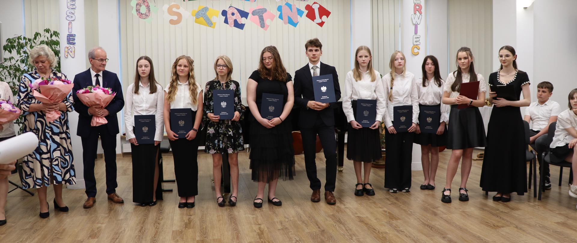 Wręczenie dyplomów ukończenia szkoły muzycznej. Na środku sali stoją nauczyciele i uczniowie szkoły. Uczniowie w rękach trzymają dyplomy. Nauczyciele bukiety kwiatów.