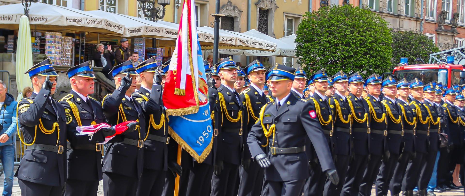 Zdjęcie przedstawia funkcjonariuszy PSP podczas Wojewódzkich Obchodów Dnia Strażaka w Gdańsku, Strażacy urani w mundury galowe, ustawieni w szeregu razem ze sztandarem