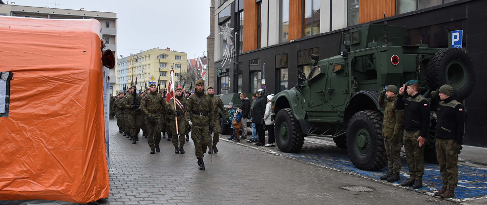 Na zdjęciu widoczna jest grupa żołnierzy Wojska Polskiego maszerujących ulicą w szyku podczas defilady. Po prawej stronie stoi wojskowy pojazd opancerzony w kolorze zielonym, obok którego stoi kilku żołnierzy w mundurach, salutujących. Po lewej stronie kadru znajduje się pomarańczowy namiot. W tle widoczne są budynki mieszkalne i przechodnie obserwujący defiladę. Scena ma miejsce na miejskiej ulicy, podczas oficjalnego wydarzenia wojskowego.