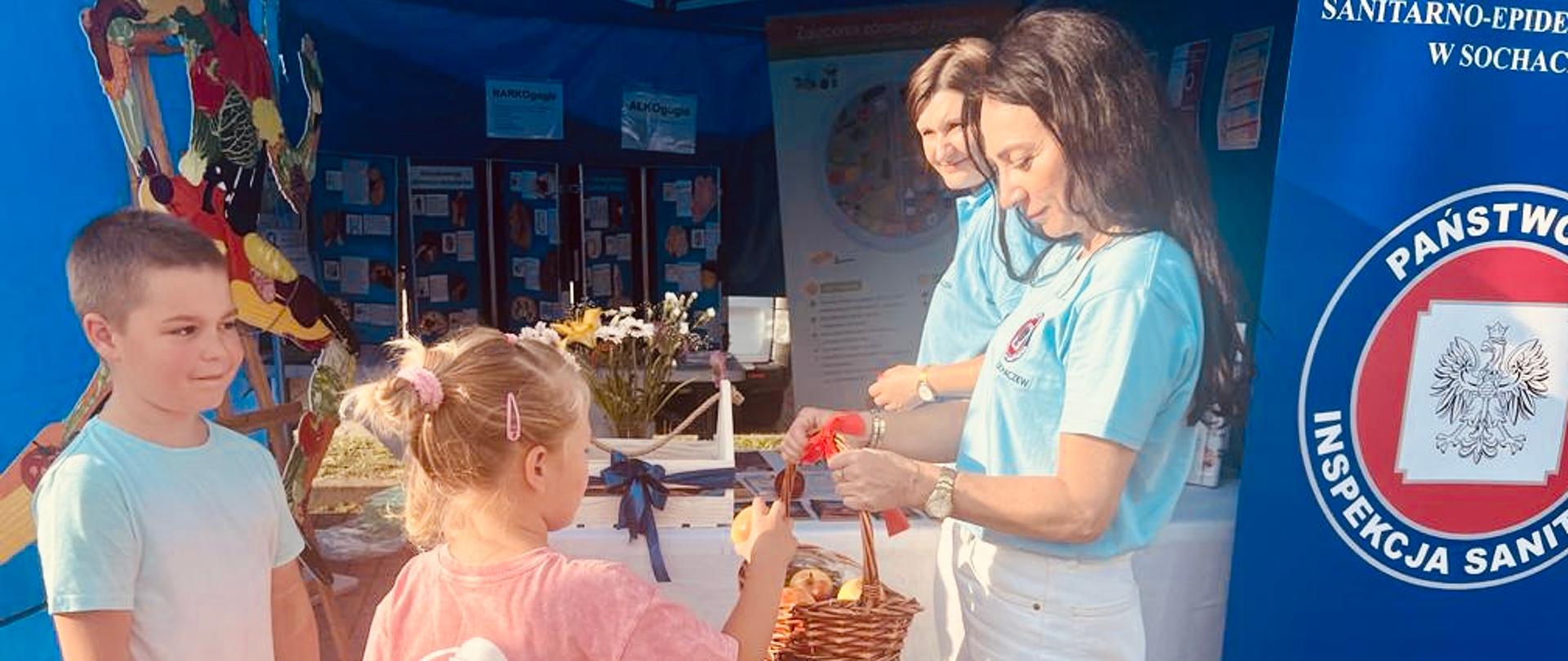 Pracownik PSSE w Sochaczewie częstuje dzieci jabłkami przed stoiskiem sanepidu.
