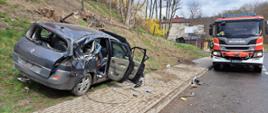 Samochód osobowy rozbity na poboczu jezdni, oparty o słup komunikacyjny który w wyniku wypadku się złamał, w tle samochód pożarniczy