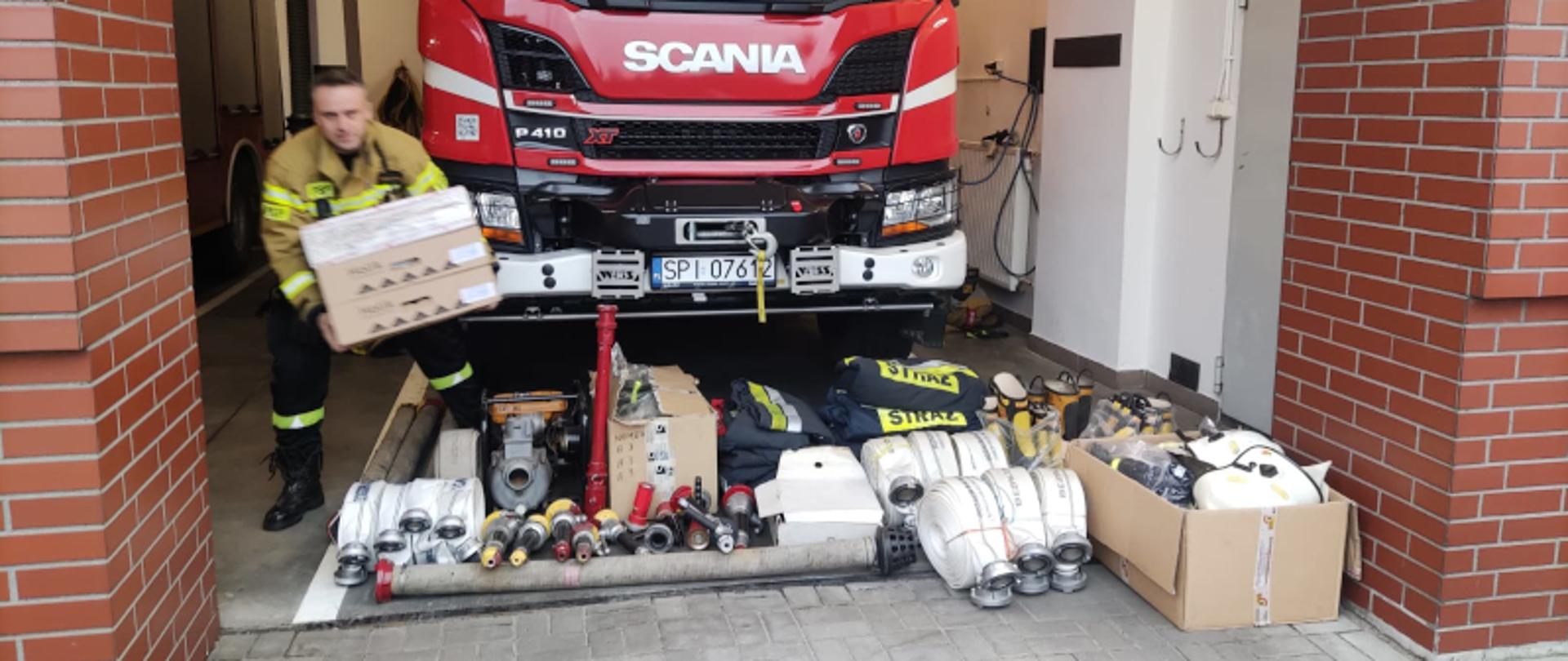 Na zdjęciu samochód ratowniczo-gaśniczy, przed samochodem zmagazynowany sprzęt pożarniczy dla Ukrainy oraz strażak przygotowujący sprzęt do przekazania 