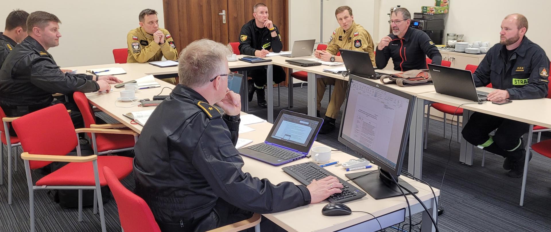 Na zdjęciu widać 8 strażaków państwowej straży pożarnej siedzących przy stole wraz z zastępcą dyrektora Biura Planowania Operacyjnego KG PSP. Na stole monitory i laptopy.