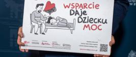Mężczyzna w policyjnym mundurze trzyma w dłoniach prostokątną białą planszę edukacyjną. Na planszy znajduje się ilustracja przedstawiająca dorosłego wspierającego leżące dziecko oraz napis „Wsparcie daje dziecku moc”. W dolnym rogu tabliczki widnieją logotypy organizatorów oraz kod QR.