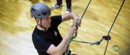 Mężczyzna w kasku ochronnym i czarnym stroju z logotypem straży wykonuje ćwiczenia z zakresu technik linowych w hali sportowej. Zawieszony na linie, koncentruje się na wiązaniu węzła. W tle widoczna drewniana podłoga i nogi innego uczestnika.