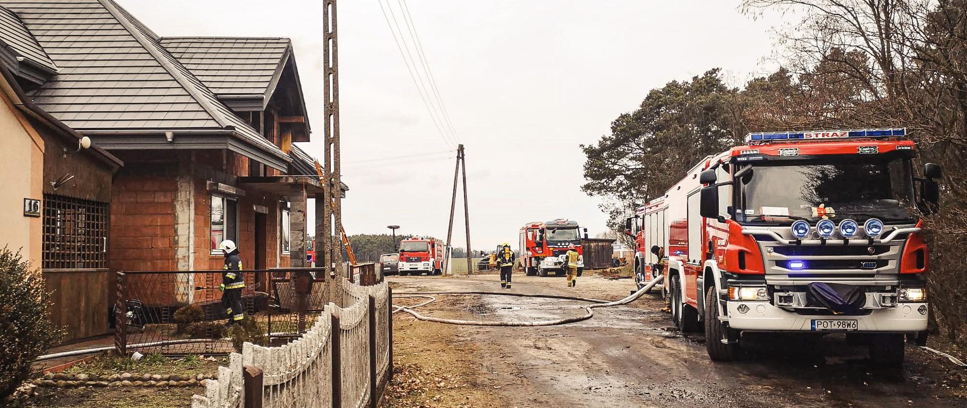 Po lewej stronie palące się obiekty, po prawej stronie pojazdy strażackie ustawione na drodze gruntowej. Za samochodami las.