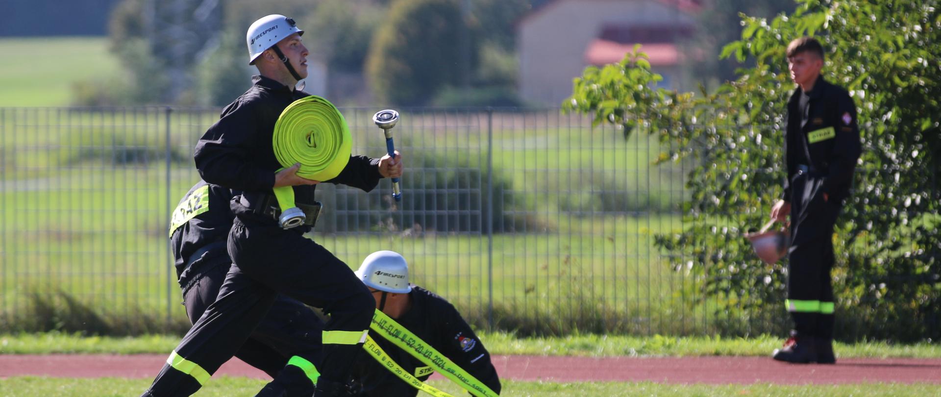Zaciekła akcja drużyny strażackiej w zawodach sportowo-pożarniczych, gdzie jeden z uczestników biegnie ze zwiniętym wężem i prądownicą, podczas gdy inni sprawiają linię główną.