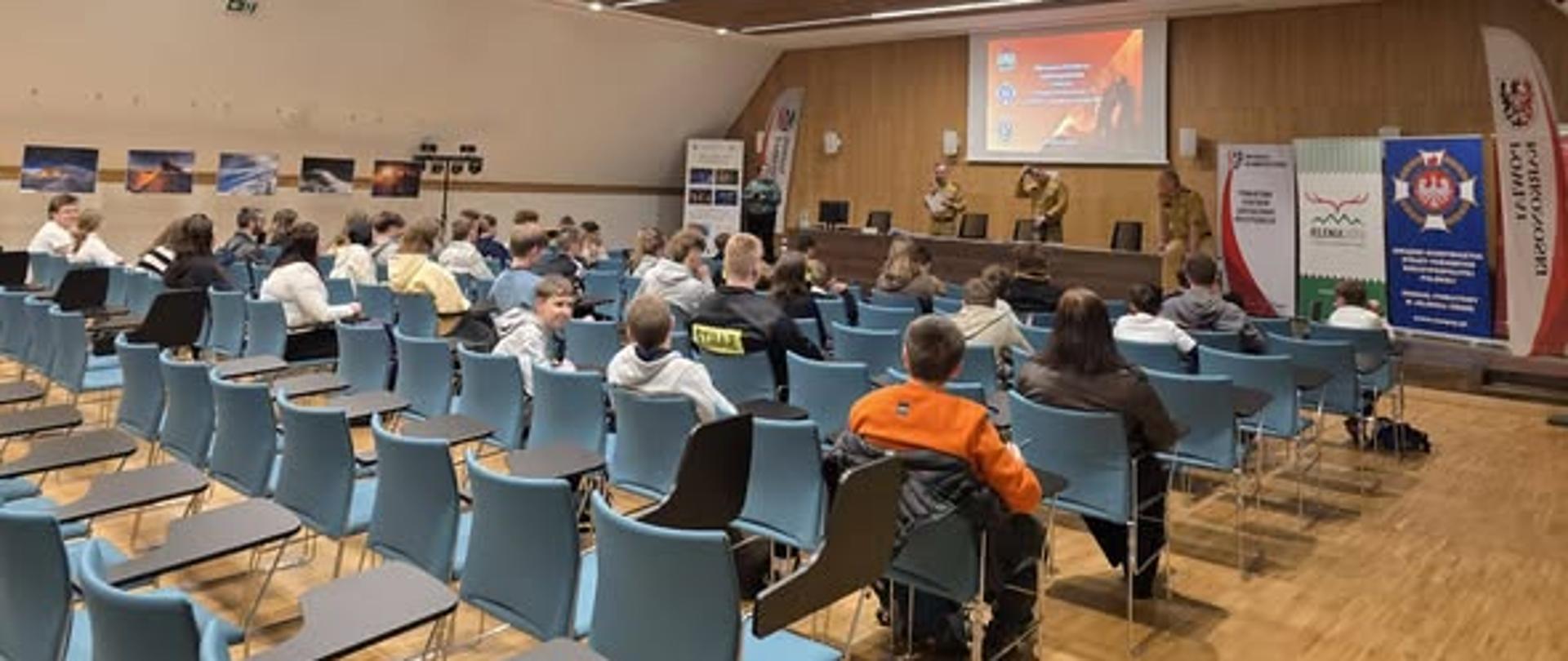 Na zdjęciu widać salę konferencyjną z rzędami krzeseł skierowanych w stronę sceny. Na krzesłach siedzi publiczność, a część miejsc jest pusta. Z przodu znajdują się osoby w mundurach stojące przy mównicy oraz ekran projekcyjny z wyświetlaną prezentacją. Po bokach sceny ustawione są roll-upy i flagi. Wnętrze jest nowoczesne, z drewnianym sufitem i jasnymi ścianami, a atmosfera przypomina oficjalne spotkanie, wykład lub konferencję.