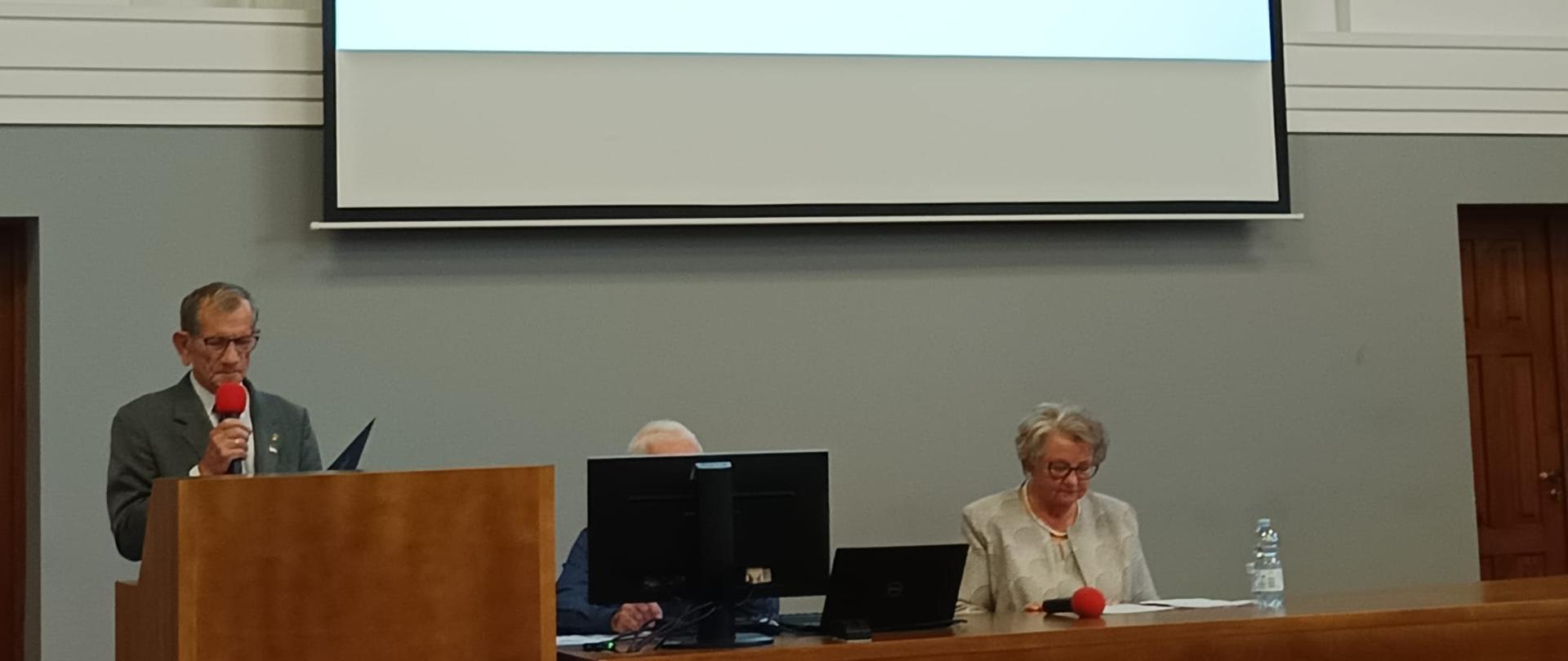 Mężczyzna w szarym garniturze, Pełnomocnik Wojewody, stoi przy mównicy odczytując list od Wojewody na tle wyświetlanej prezentacji 