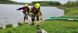 Dwóch strażaków zabezpieczonych w kamizelki asekuracyjne sprawia linii ssawną w tle woda oraz drzewa