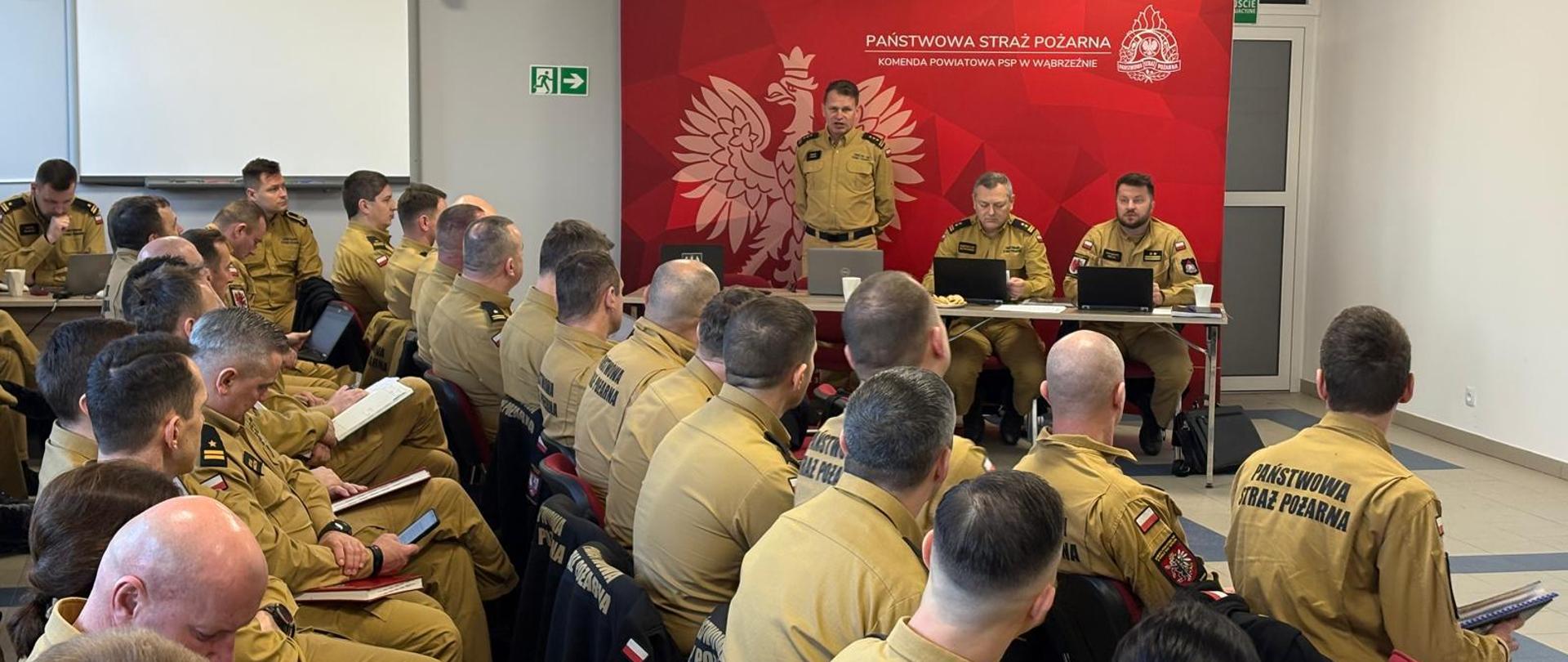 Na sali, w szeregach siedzą strażacy - kilkadziesiąt osób, w mundurach służbowych. W dali stół prezydialny, przy nim trzech strażaków, jeden stoi i przemawia, dwaj pozostali patrzą w komputery. Za nimi ścianka firmowa, w kolorze czerwonym. 