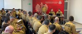 Na sali, w szeregach siedzą strażacy - kilkadziesiąt osób, w mundurach służbowych. W dali stół prezydialny, przy nim trzech strażaków, jeden stoi i przemawia, dwaj pozostali patrzą w komputery. Za nimi ścianka firmowa, w kolorze czerwonym. 