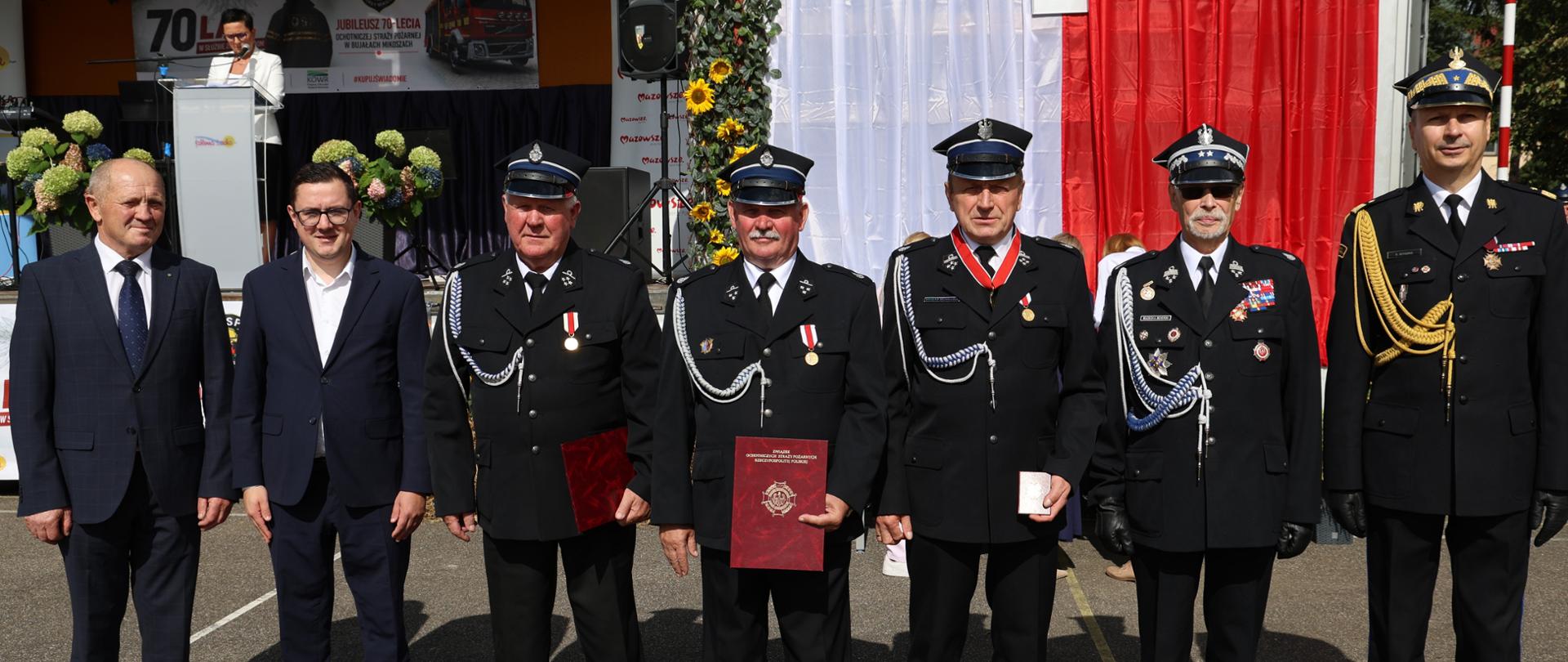 Pamiątkowe zdjęcie na tle sceny z banerem jubileeuszowym „Jubileusz 70-lecia Ochotniczej Straży Pożarnej w Bujałach Mikoszach”.
Przedstawia odznaczonych druhów oraz przedstawicieli władz i PSP trzymających dyplomy, legitymacje oraz medale.
Po lewej stronie na scenie widoczna jest spikerka prowadząca uroczystość przy mównicy.