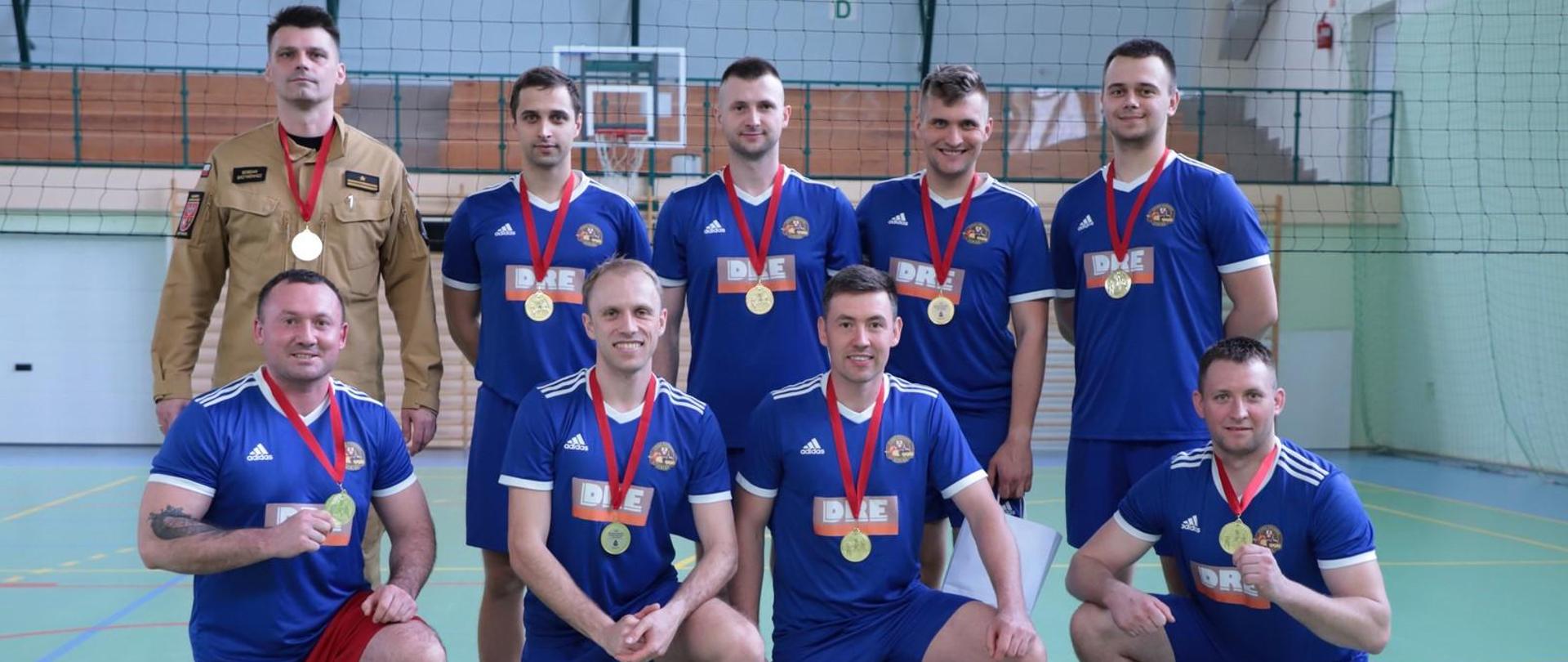 Zwycięska drużyna z Elbląga pozuje do zdjęcia w strojach sportowych wraz z komendantem miejskim. Zawodnicy mają na szyi zawieszone złote medale, a przed nimi stoją torby z upominkami.