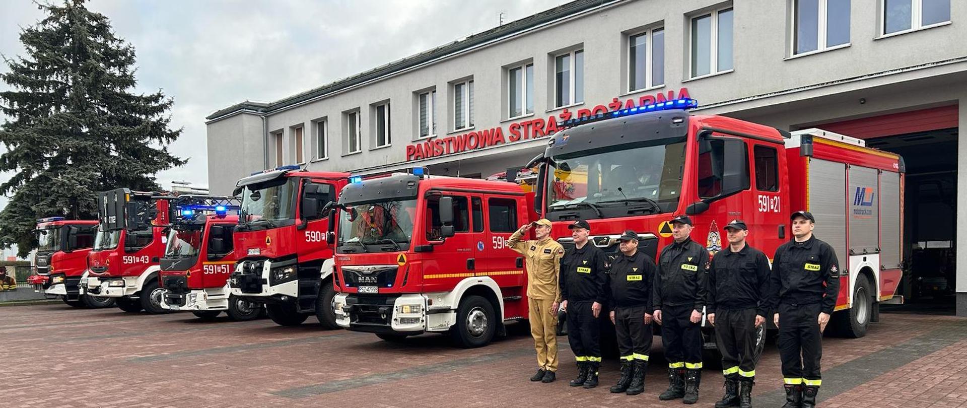 Strażacy przy samochodzie przed budynkiem komendy